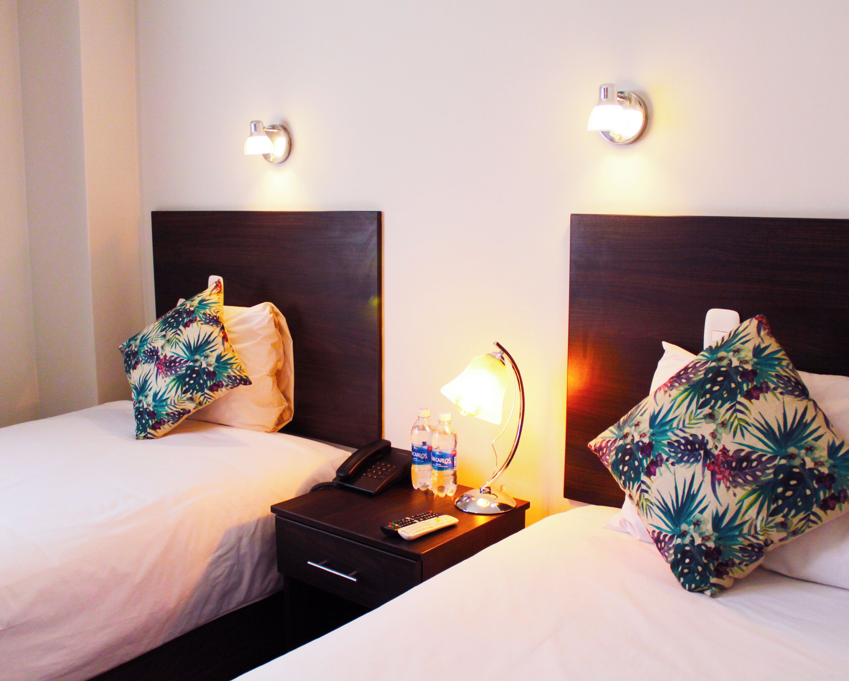 Habitación de hotel con dos camas, cada una con un cojín floral y una mesita de noche con lámpara, teléfono y botellas de agua.