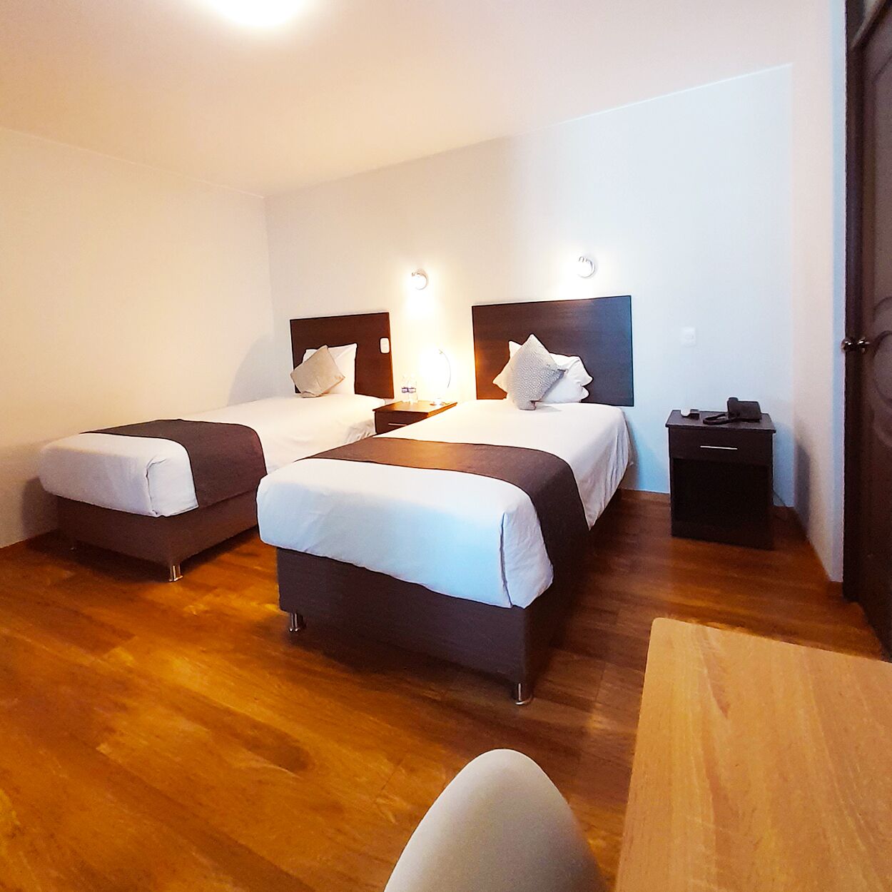 Quarto de hotel com duas camas, piso de madeira, uma escrivaninha e uma cadeira.