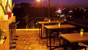 Un área de comedor al aire libre con mesas, sillas, plantas en macetas y luces colgantes.