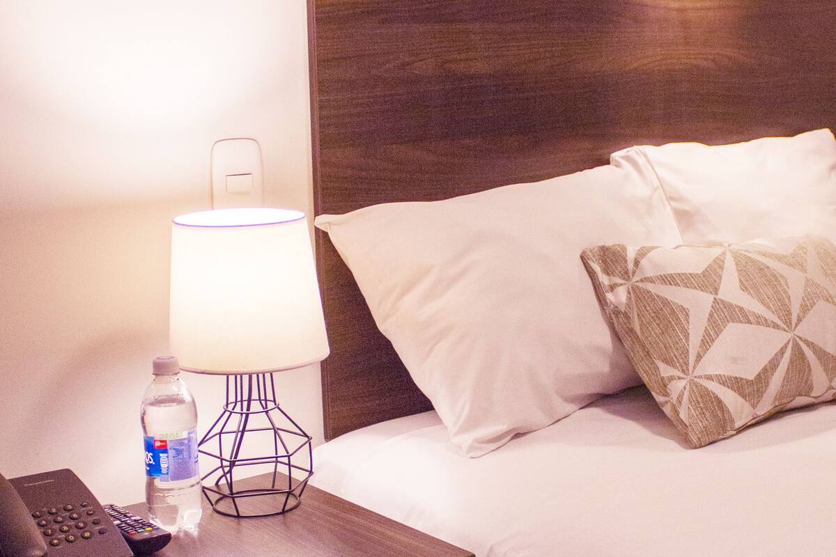 Una habitación de hotel con una cama, almohadas, una lámpara, una botella de agua y un control remoto sobre una mesita de noche de madera.