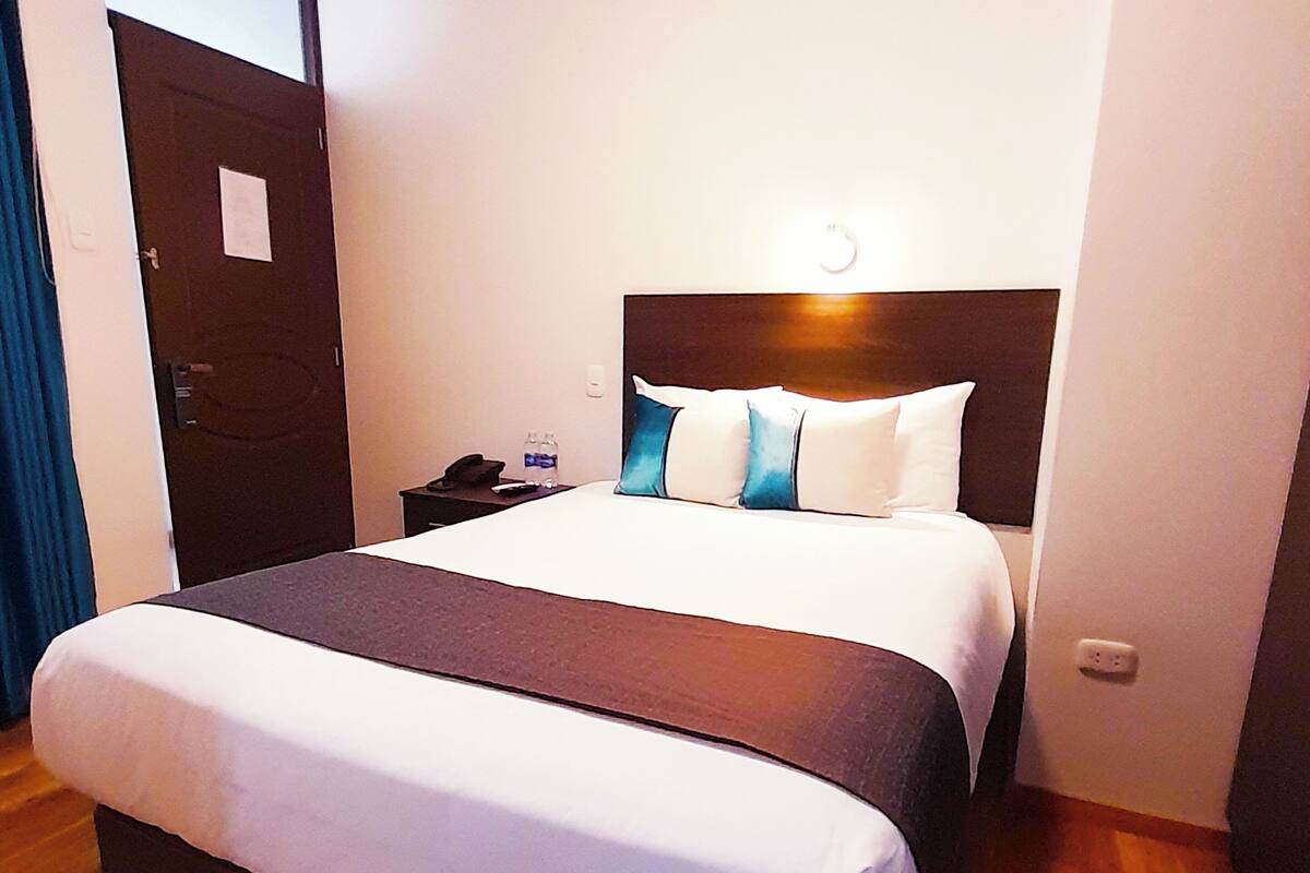 Habitación de hotel con una cama grande, una mesita de noche con teléfono y botella de agua, y una puerta con un cartel.