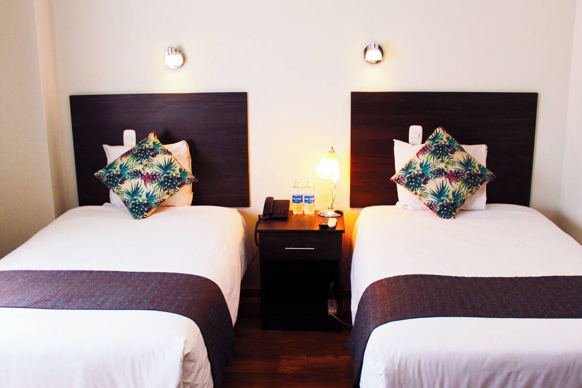 Habitación de hotel con dos camas, cada una con cabecero oscuro y almohada estampada con motivos florales. Entre las camas hay una mesita de noche con una lámpara, un teléfono y una botella de agua.