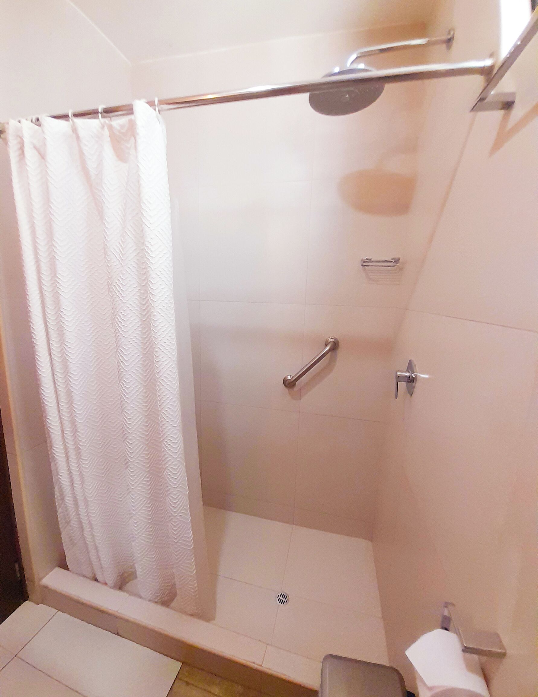 Un área de ducha con cortina blanca, un cabezal de ducha manual, un control manual para la ducha y un pequeño cubo de basura.