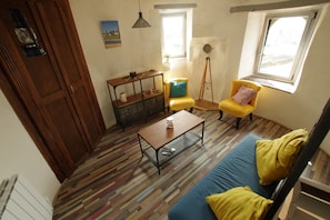 A room with a wooden door, a blue sofa with yellow cushions, a wooden coffee table, a small side table, a floor lamp, and a window with a view.