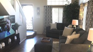 A living room with a sofa, ottoman, and a television displaying a man. There is a staircase in the background, a window with blinds, and a potted plant. - Beautiful 2 BR Plus Loft, Attached 2 Car Garage, Recently Updated! (Scottsdale)