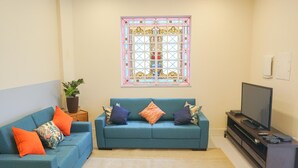 A living room with a teal sofa, a large window with stained glass, a television, and a small plant. - Quintal Hostel (São Paulo)