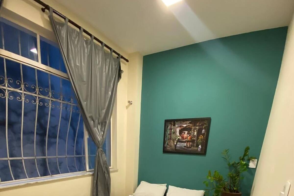 A bedroom with a bed, a window with curtains, a nightstand with a plant, and a wall-mounted picture.