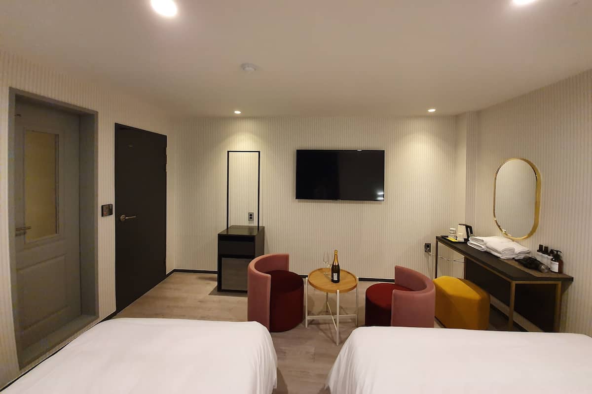 A hotel room with two beds, a TV, a mirror, a small table with a bottle and glass, and a desk with toiletries.