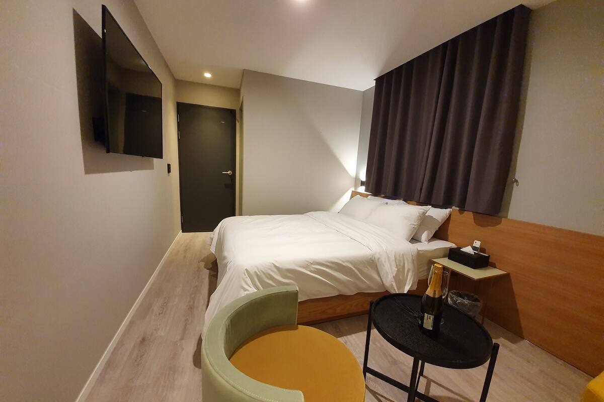 A hotel room with a bed, a small table with a bottle, a chair, and a TV mounted on the wall.