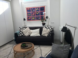 Uma sala de estar moderna com um sofá preto, almofadas estampadas, uma mesa de centro redonda de vime, uma planta em vaso e uma obra de arte emoldurada na parede.