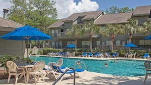 A pool area with lounge chairs and umbrellas, surrounded by buildings with balconies and palm trees. - Egret Point - Large 3BR/3BA - Great Location with Water View and Pool (Hilton Head Island)