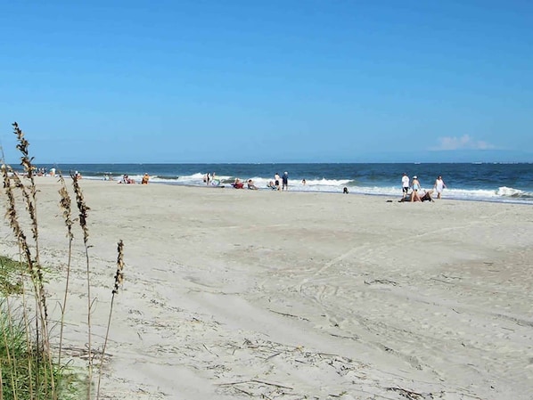 A wide sandy beach with people walking and sitting, and the ocean in the background.