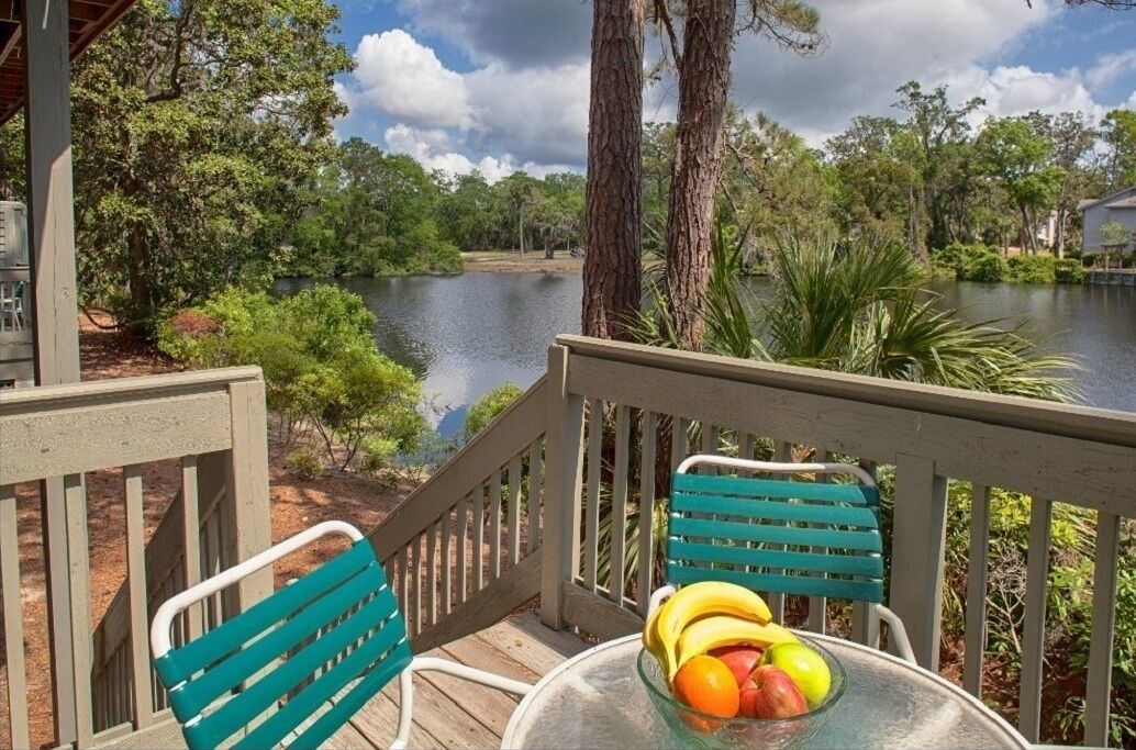 A wooden deck with two chairs and a table, overlooking a calm lake with trees and a building in the background.