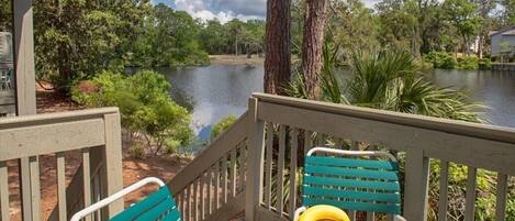 A wooden deck with two chairs and a table, overlooking a calm lake with trees and a building in the background.