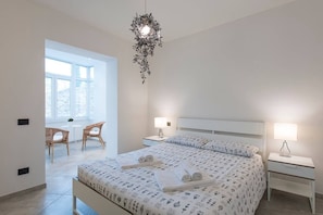 A bedroom with a large bed, two bedside tables with lamps, a hanging light fixture, a window, and a chair by the window.