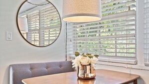 Un coin repas avec une table en bois, un banc recouvert d’un tissu gris, un miroir rond et un vase avec des fleurs blanches.