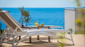 A sun lounger with a table beside it, a glass of orange juice, and a plate with fruit, overlooking a clear blue sea.