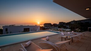 A rooftop pool with lounge chairs and a table, overlooking a sunset over the sea and a residential area.