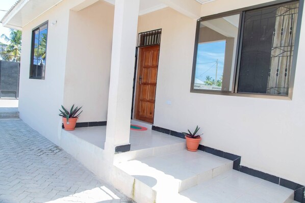 Una casa con fachada blanca, puerta de madera y dos ventanas. Hay escalones que conducen a la puerta y dos macetas con plantas en los escalones.