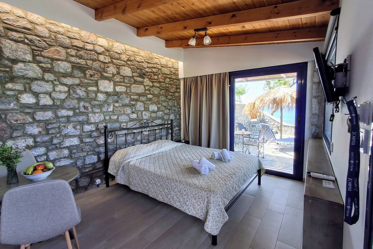 Une chambre avec un mur en pierre, un lit recouvert d’une courtepointe, une petite table avec des fruits, une chaise et une vue sur un espace extérieur avec une piscine et des parasols.