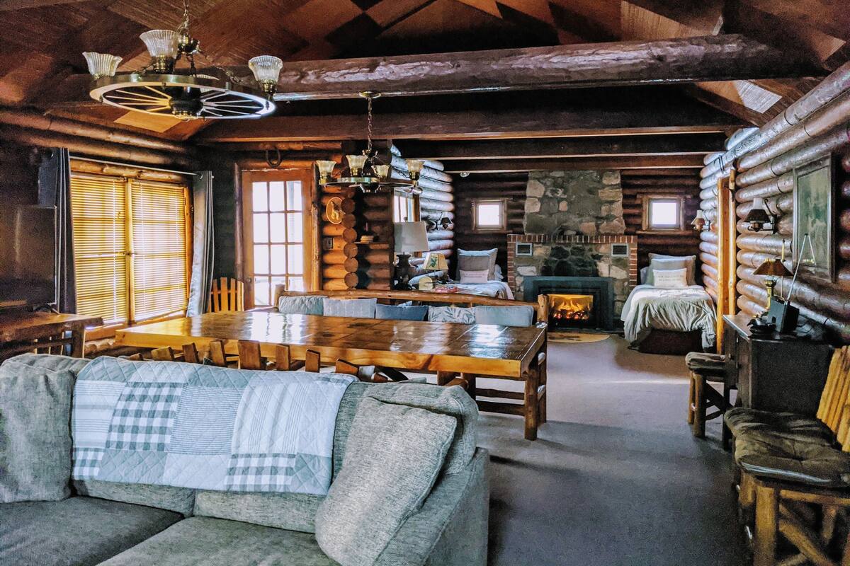 Interior de cabana de madeira com área de jantar, sofá, lareira e parede de pedra.
