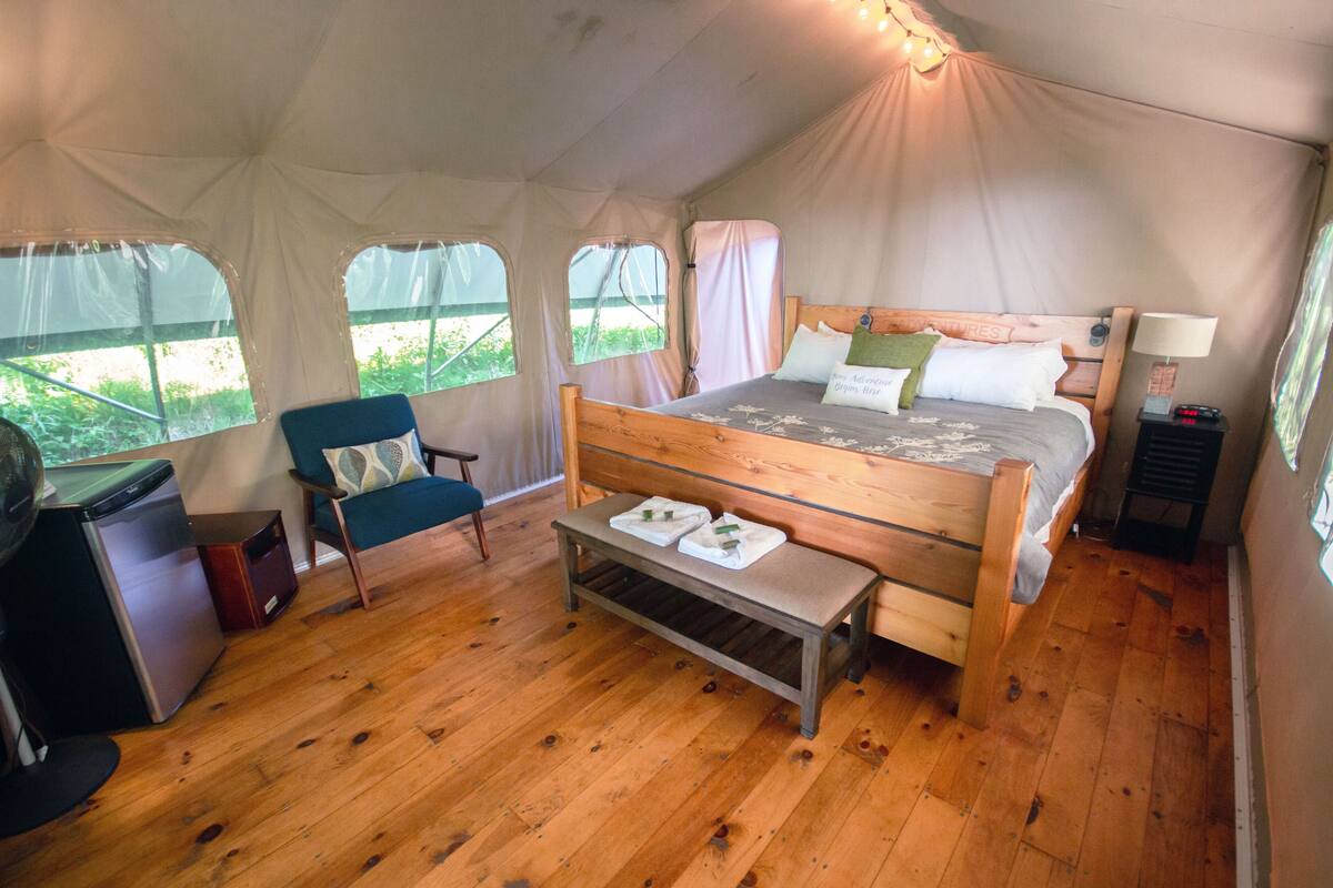 Acomodação em tenda com uma cama de madeira, uma cadeira, uma mesinha e uma escrivaninha com geladeira.