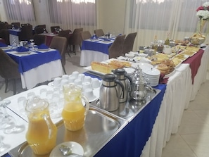 Montaje tipo bufé con diversos alimentos, estaciones de café y té, y mesas ordenadas con manteles azules.