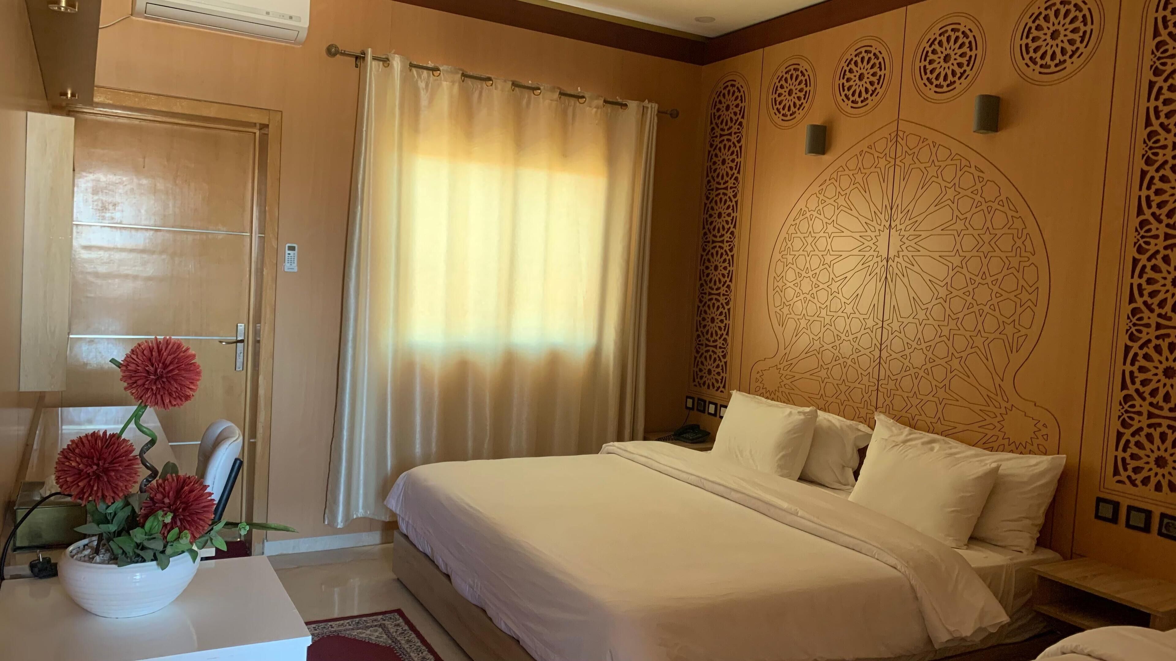 A hotel room with a bed, a nightstand, a vase with flowers, a curtain, and a wall with intricate patterns.
