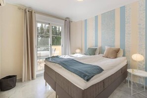 Ein Schlafzimmer mit einem Bett, einem Fenster mit Vorhängen, einem kleinen Tisch mit Lampe und einer Wand mit vertikalen Streifen.