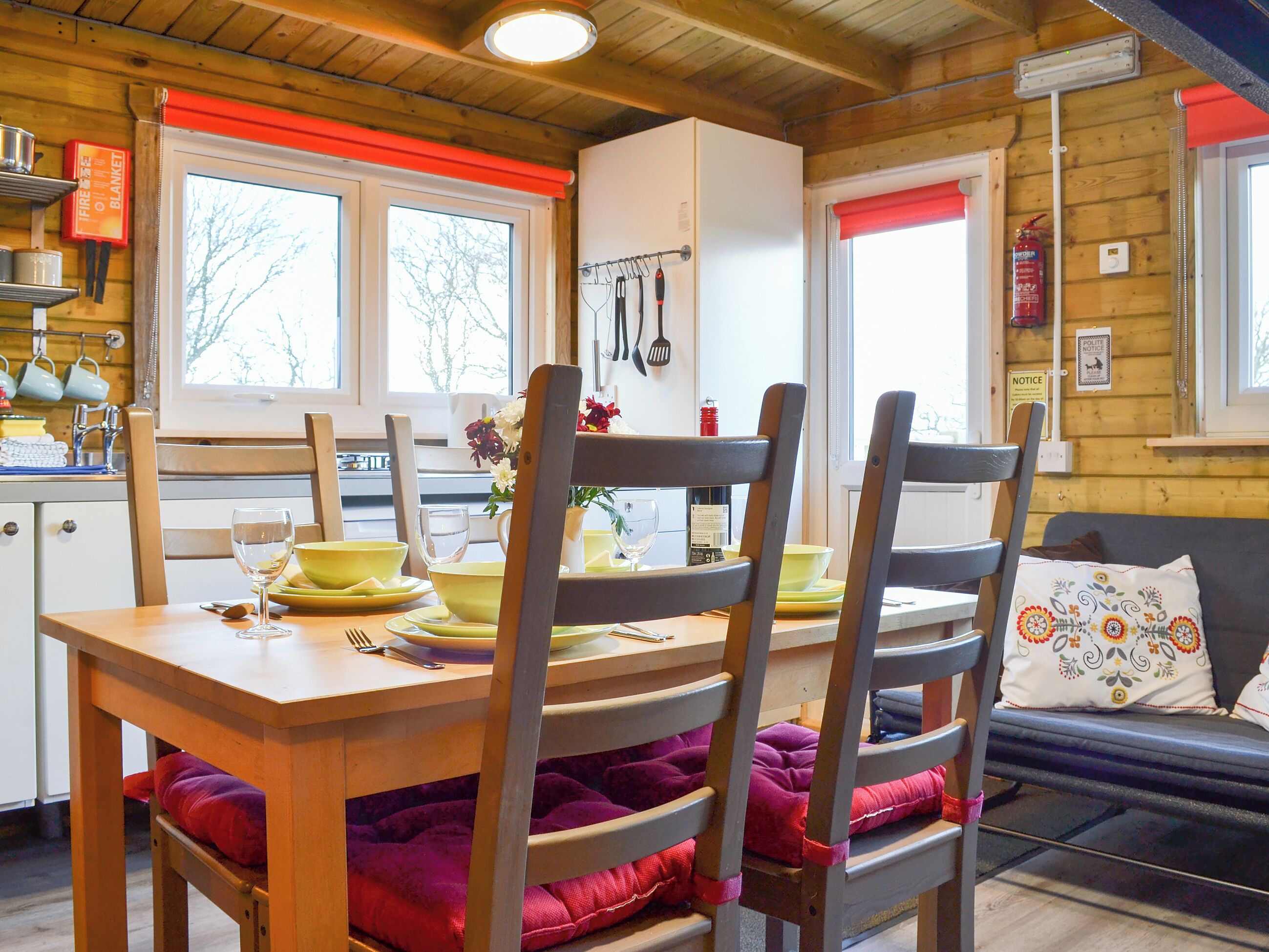 A cozy cabin-style kitchen with wooden walls, a dining table set with yellow plates and a vase of flowers, and a red blanket on the chair cushions.