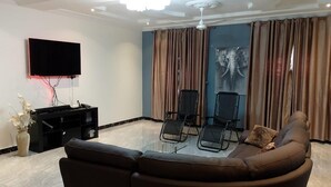 Een woonkamer met een bruine bank, twee zwarte stoelen, een flatscreen televisie, een plafondventilator en een groot raam met gordijnen.