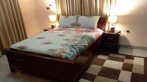 Een net opgemaakt bed met een bedsprei met patroon, twee bedlampen, een klein houten nachtkastje en een geruit vloerkleed.