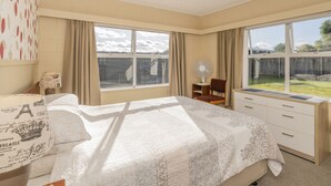A bedroom with a bed, a desk, and a window with a view of a backyard. - Lintons Sunshine Retreat - Whangamata Home (Whangamata)