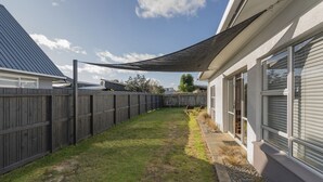 A backyard with a wooden fence, a shaded area, and a house with large windows. - Lintons Sunshine Retreat - Whangamata Home (Whangamata)