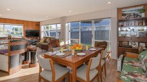 A spacious living area with a wooden dining table, comfortable seating, a TV, and a bookshelf. - Lintons Sunshine Retreat - Whangamata Home (Whangamata)