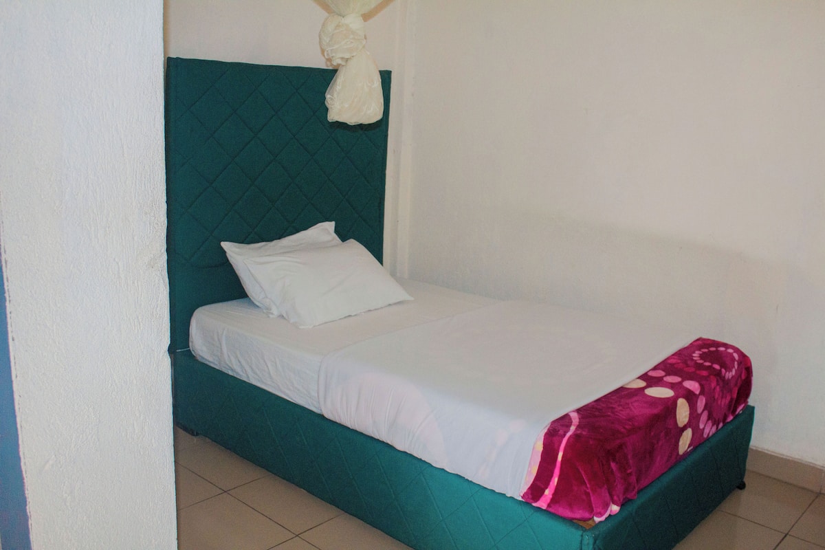 Una cama individual con cabecera color turquesa y ropa de cama blanca, una almohada blanca y una manta rosa con lunares.