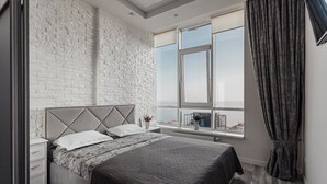 A modern bedroom with a brick wall, a large window with curtains, a bed with a grey quilt, and a nightstand with a vase of flowers. - Apartments Arcadia. Panorama of the sea. 3 bedrooms  (Odessa)