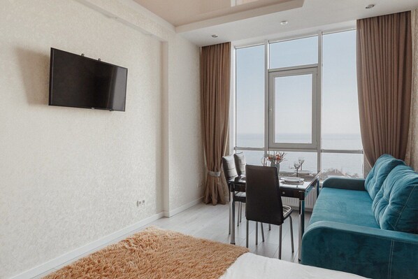 Una habitación de hotel moderna con un gran ventanal, un televisor de pantalla plana montado en la pared, una zona de comedor con mesa y sillas, y un sofá color turquesa.