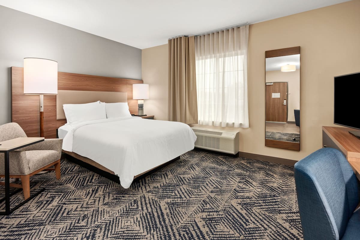 A hotel room with a large bed, two bedside lamps, a desk with a chair, a TV, a mirror, and a window with curtains.