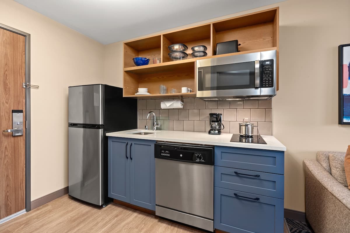 A compact kitchen with a stainless steel refrigerator, dishwasher, and microwave. There is a wooden shelf above the sink with dishes and a coffee maker.