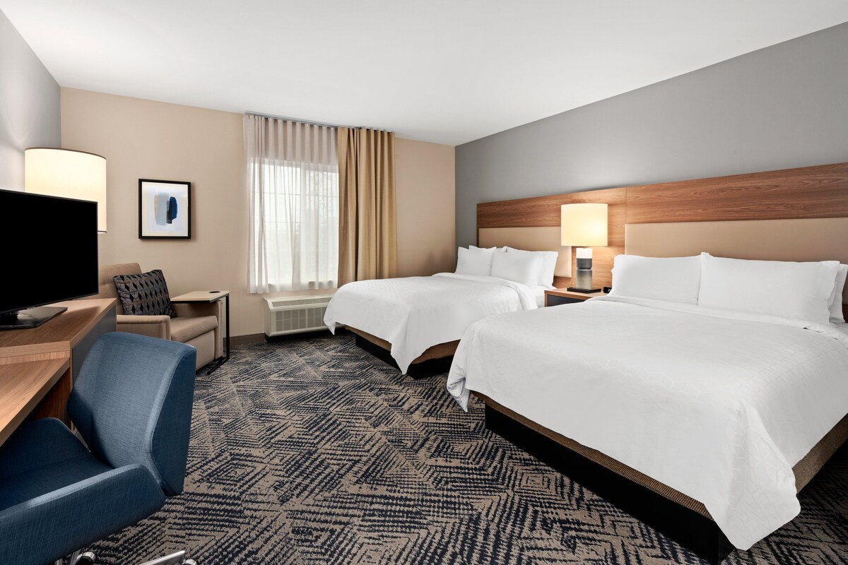 A hotel room with two beds, a desk with a chair, a television, and a large window with curtains.