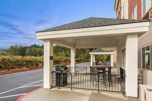 A covered outdoor seating area with a grill, tables, and chairs, adjacent to a building with a red roof. - Candlewood Suites Sumner - Puyallup Area by IHG (Sumner)