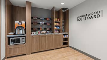 Un petit coin cuisine équipé d’une machine à café, d’étagères remplies d’en-cas et d’une pancarte indiquant « CANDLEWOOD CUPBOARD ».