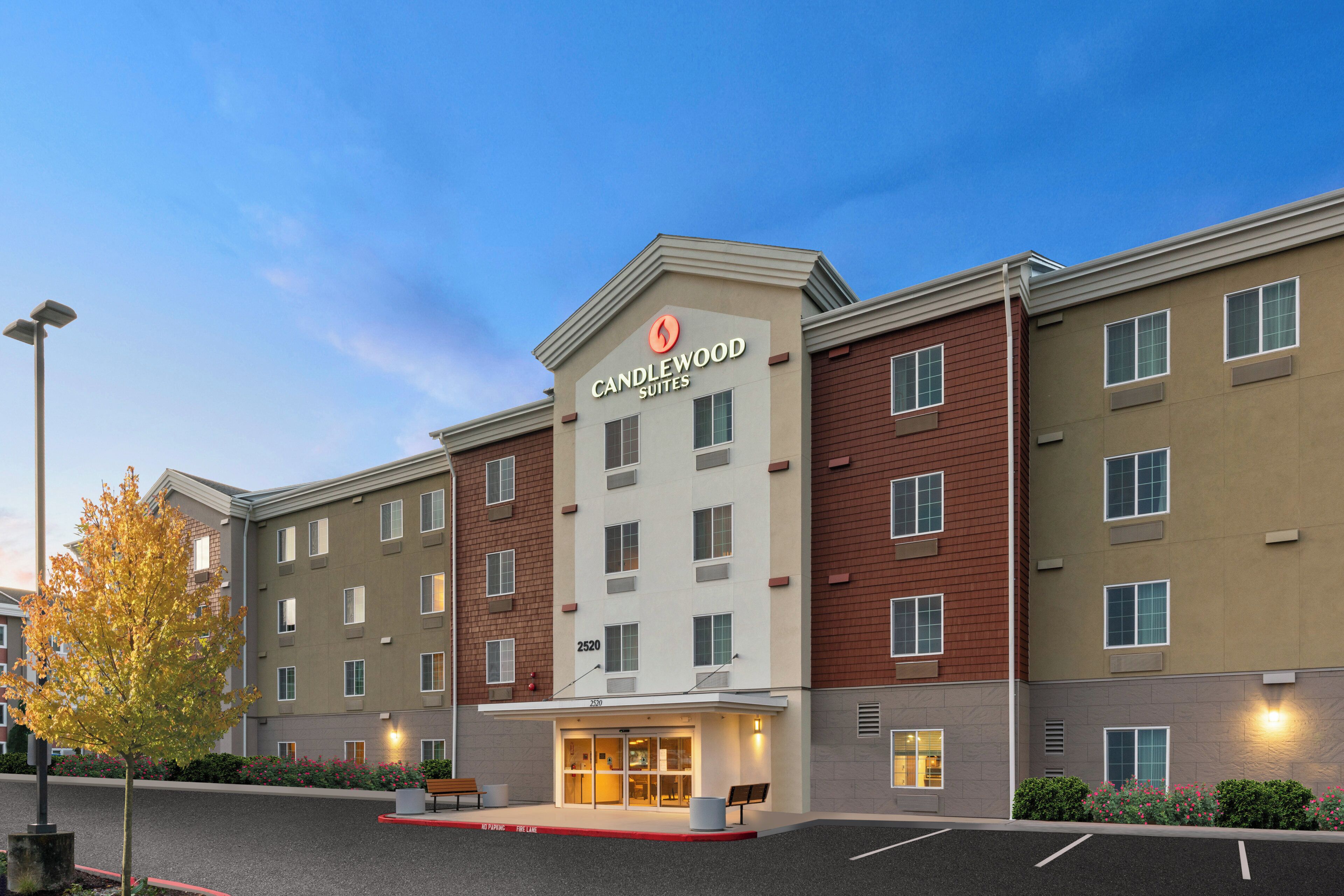 A multi-story hotel with a prominent 'Candlewood Suites' sign, a parking lot, and a landscaped area with benches.