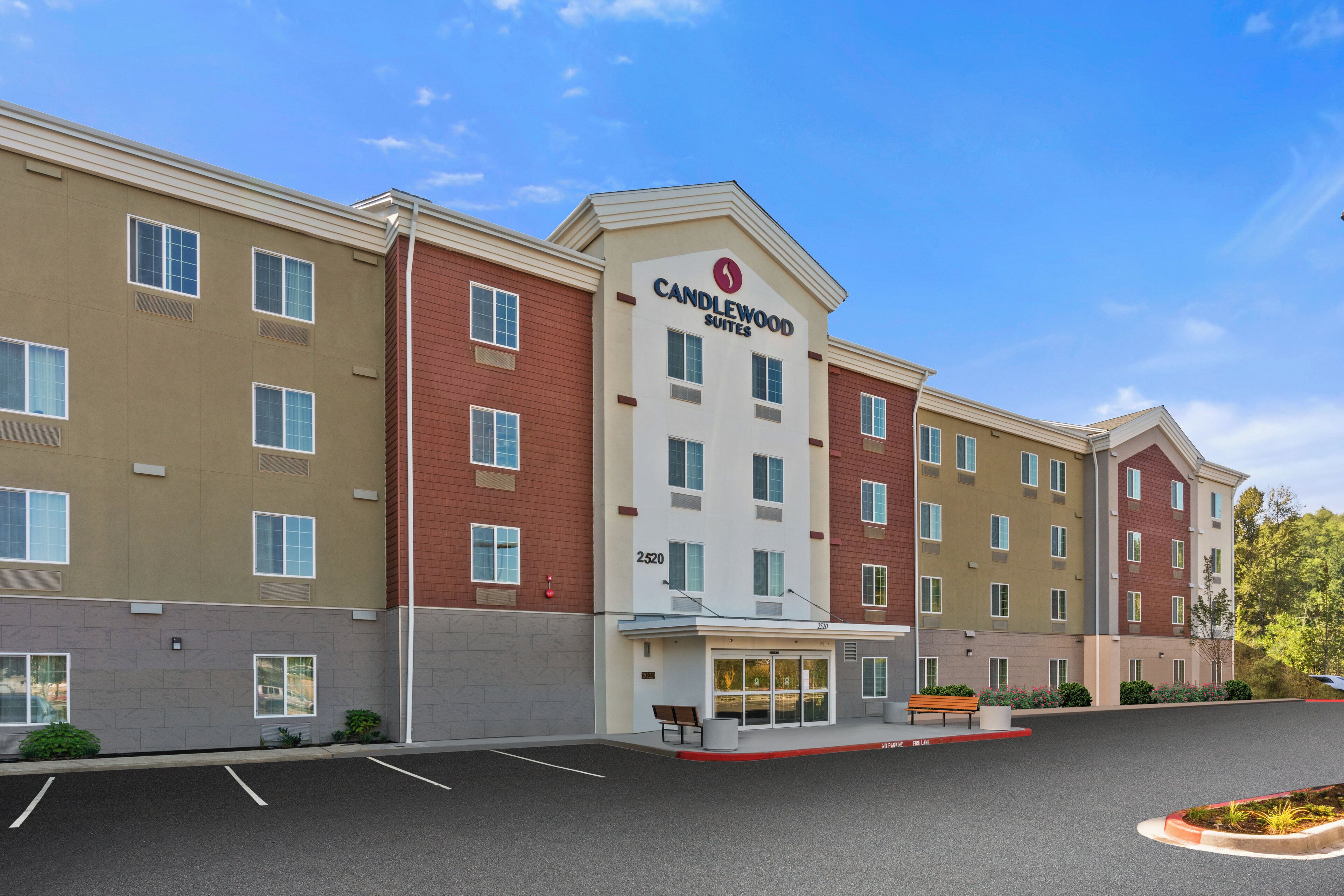 Un hotel de varios pisos con un cartel llamativo de 'Candlewood Suites', un acceso cubierto y un estacionamiento con espacios delimitados.