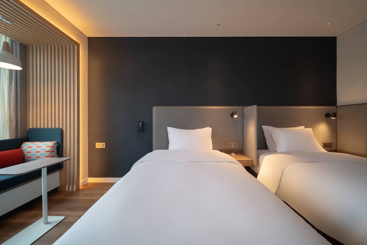 A modern hotel room with two beds, a sofa, a small table, and a window with blinds.