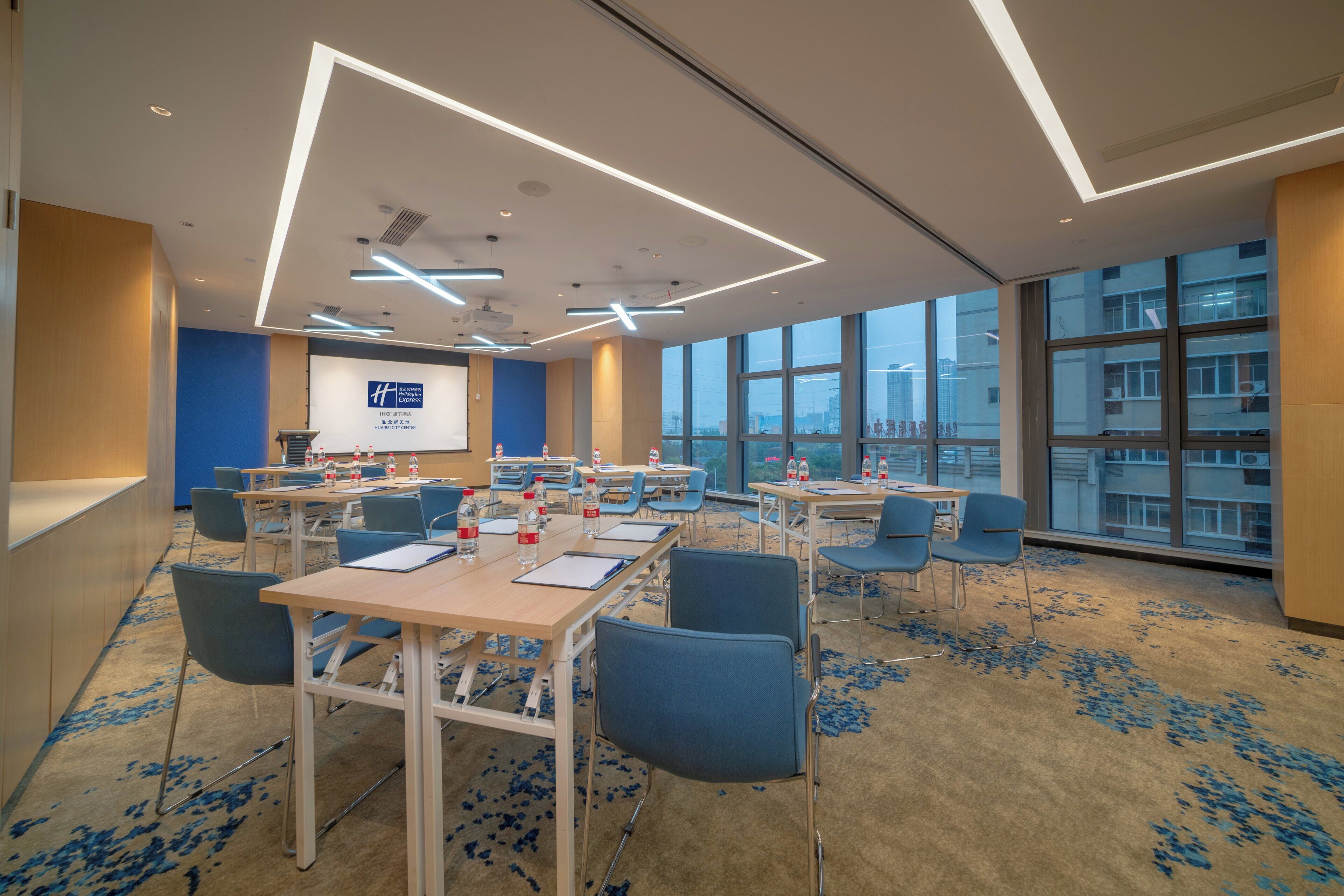 A conference room with tables, chairs, a projector screen, and a large window with a city view.