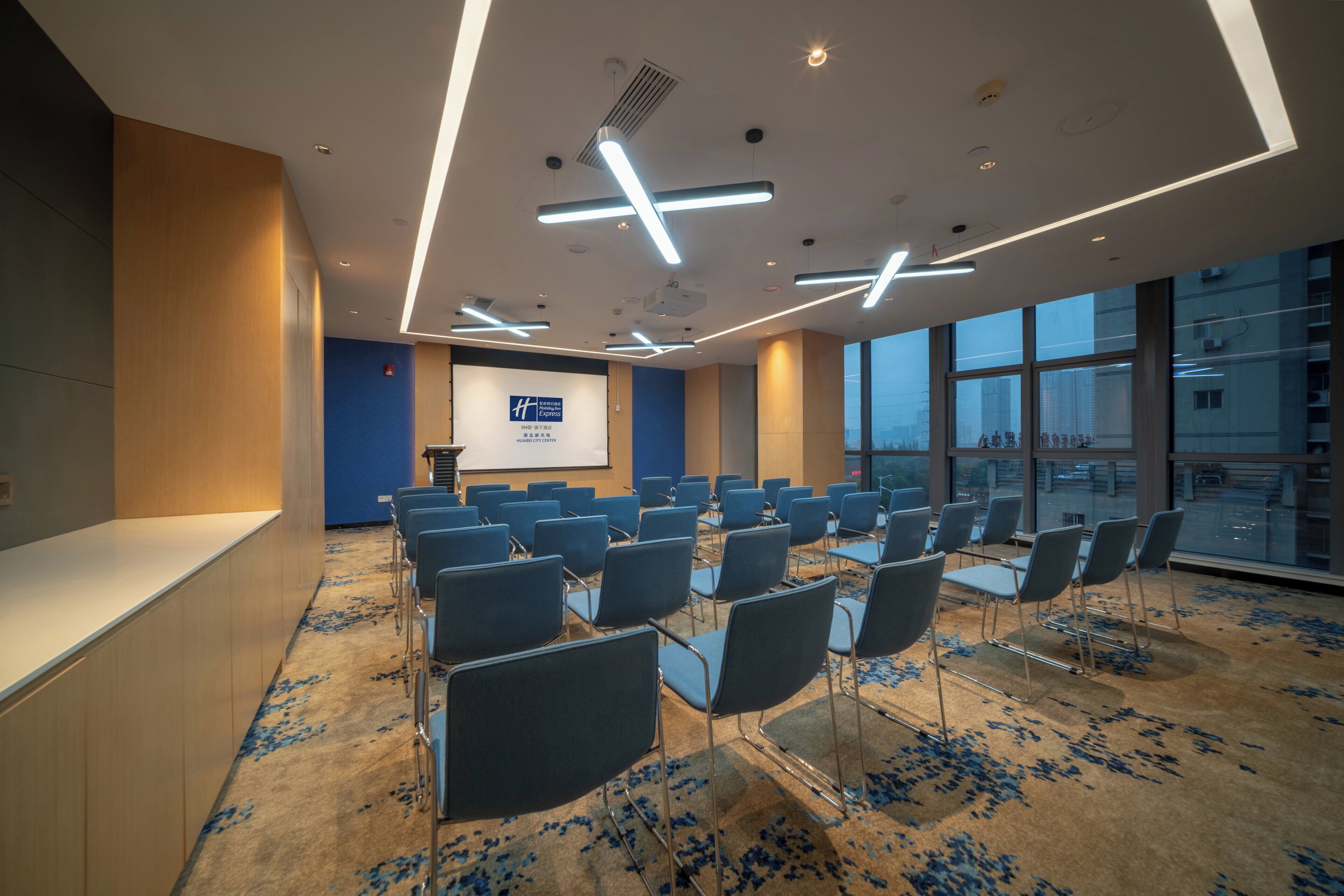 Una sala de conferencias con filas de sillas, una pantalla de presentación y ventanas amplias.