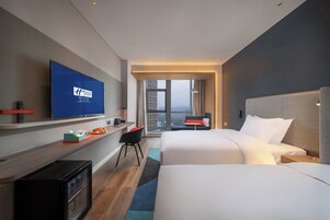 Habitación de hotel con televisor, escritorio con control remoto, cama, silla y ventana con vista a edificios.
