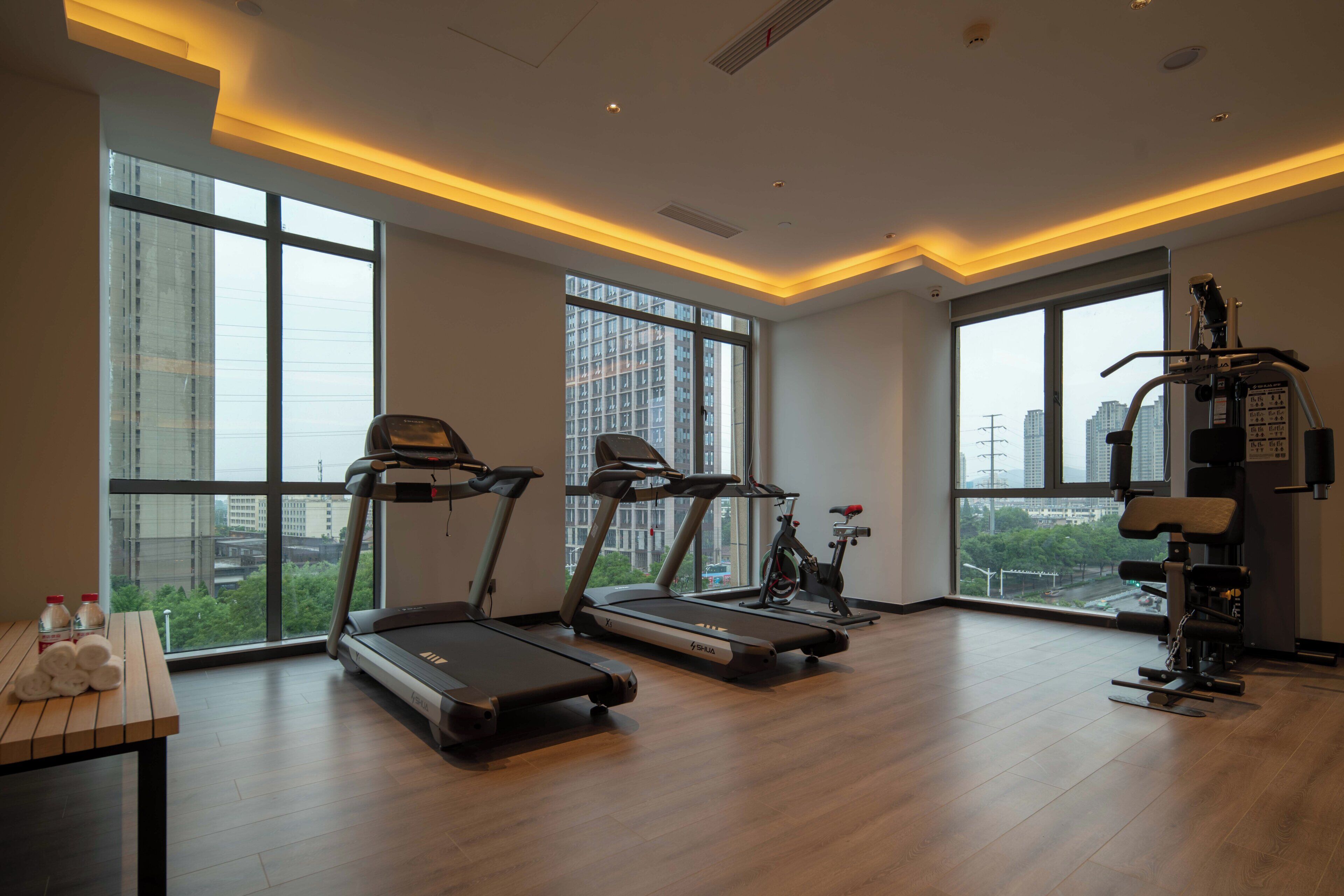A modern gym with treadmills, a stationary bike, and weight machines, featuring large windows and a high ceiling.